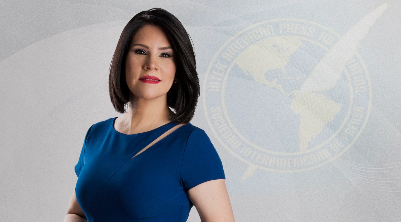 Mujer de cabello corto y vestido azul posando frente al logo de la Sociedad Interamericana de Prensa (SIP).