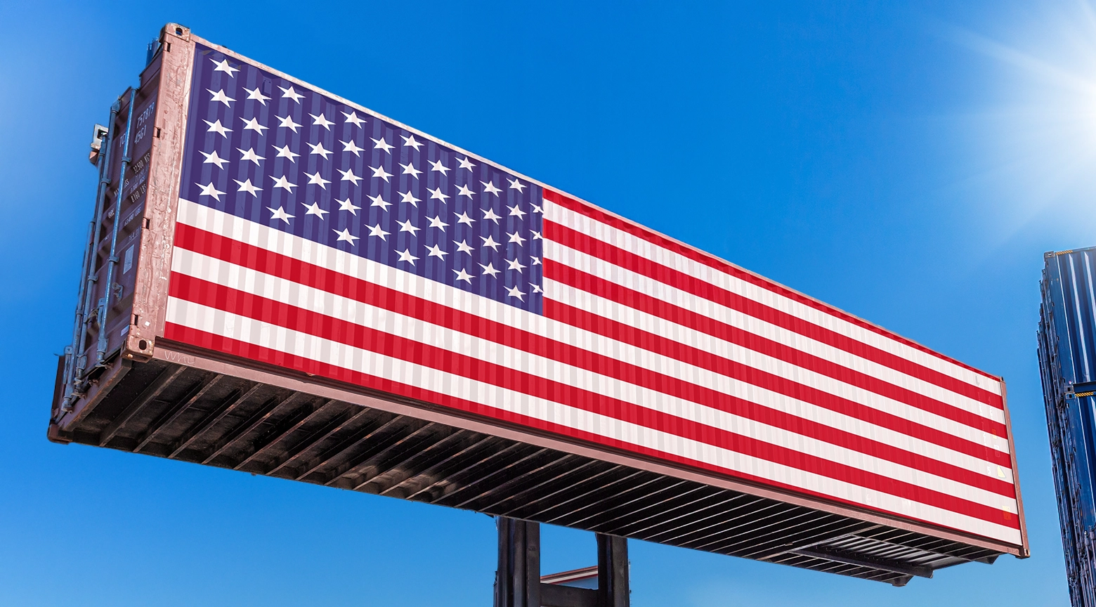 Contenedor marítimo decorado con la bandera de Estados Unidos elevado contra un cielo azul despejado.