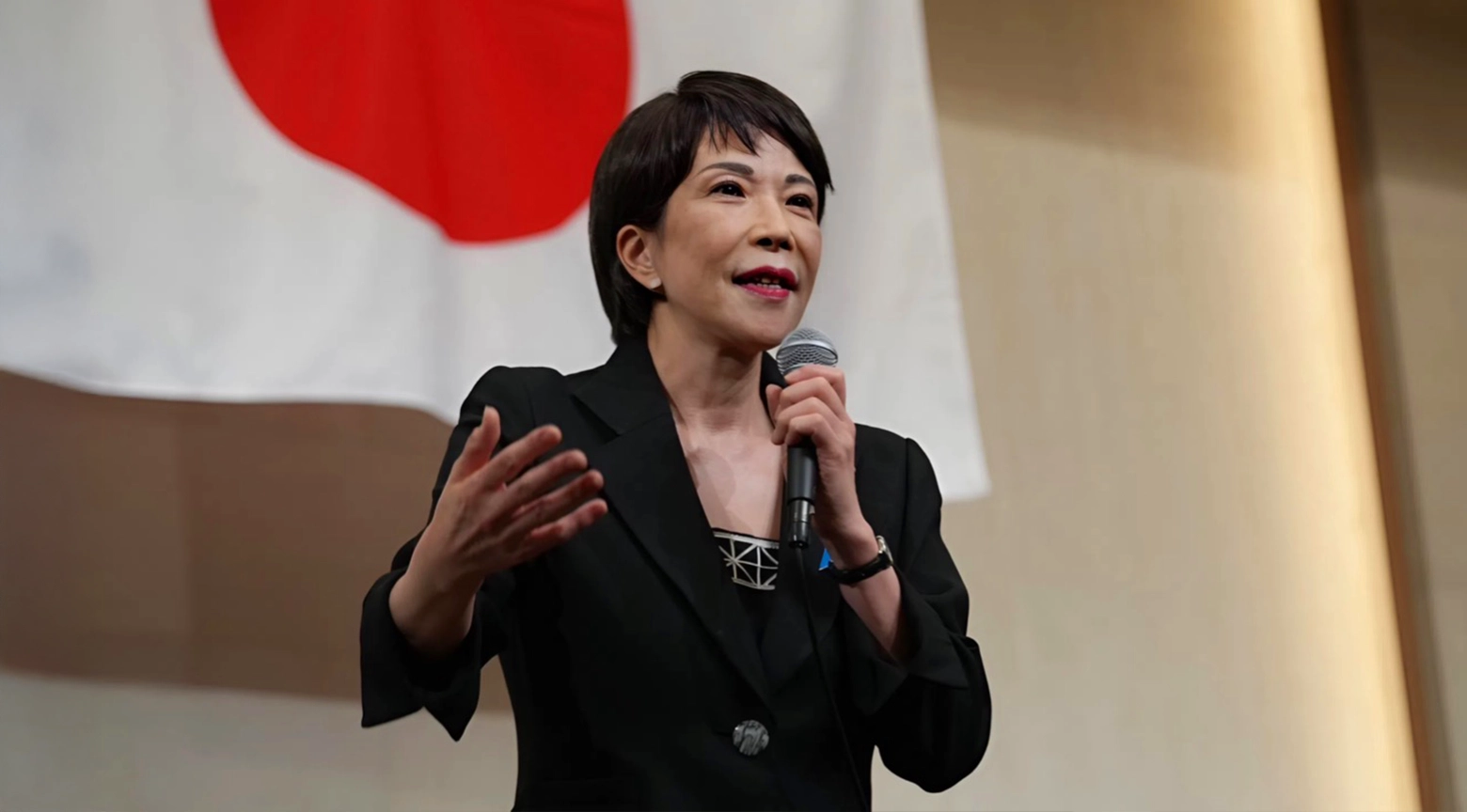 Mujer japonesa hablando con micrófono frente a una bandera de Japón durante un evento político.