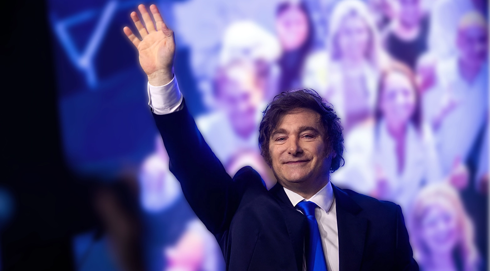 Hombre con traje azul y corbata del mismo color saluda al público con la mano levantada durante un acto político.