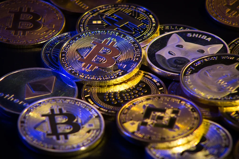 Varias monedas físicas de criptomonedas como Bitcoin, Ethereum, Shiba Inu y otras sobre una superficie oscura.