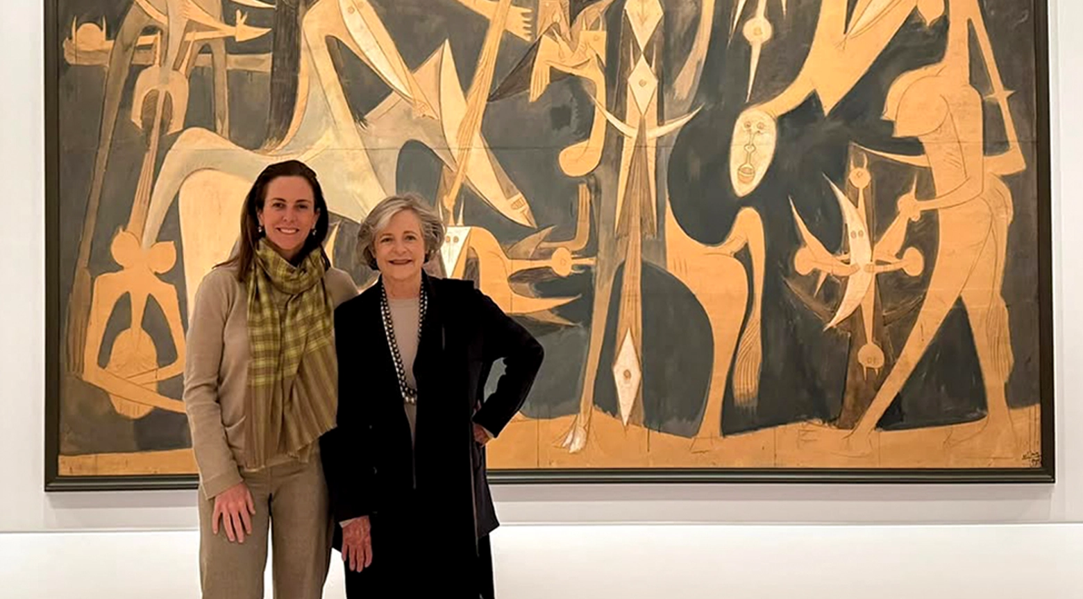 Dos mujeres posan frente a una gran pintura abstracta en tonos dorados y oscuros dentro de una galería de arte.