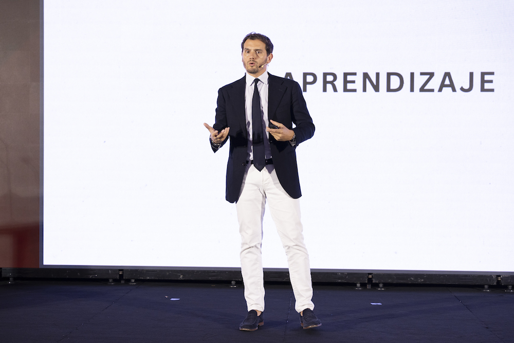 hombre de traje en un escenario dictando una conferencia con una pantalla de fondo y la palabra aprendizaje