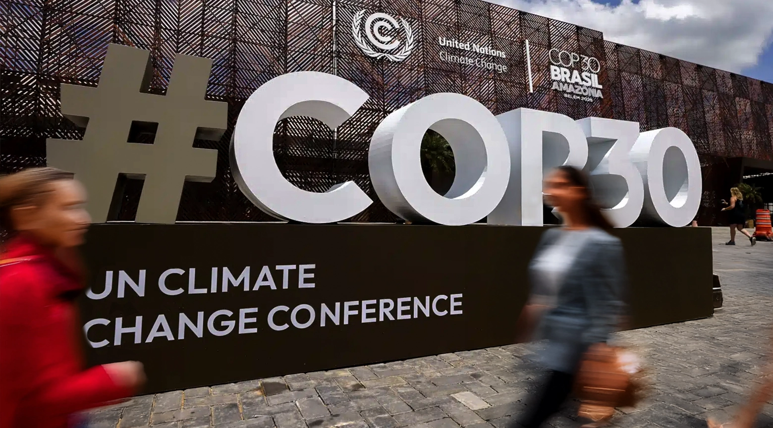 Instalación con el logotipo de la COP30 en Brasil, evento de la ONU sobre cambio climático, con personas caminando frente al cartel principal.