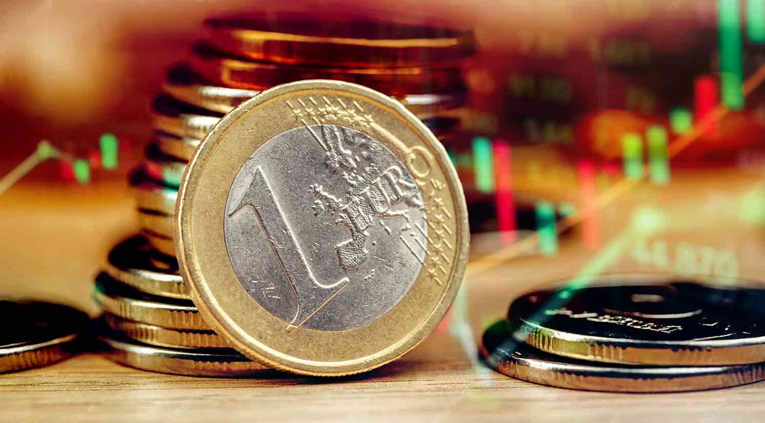 Primer plano de una moneda de un euro colocada frente a un fondo borroso con gráficos financieros en tonos rojos y verdes.