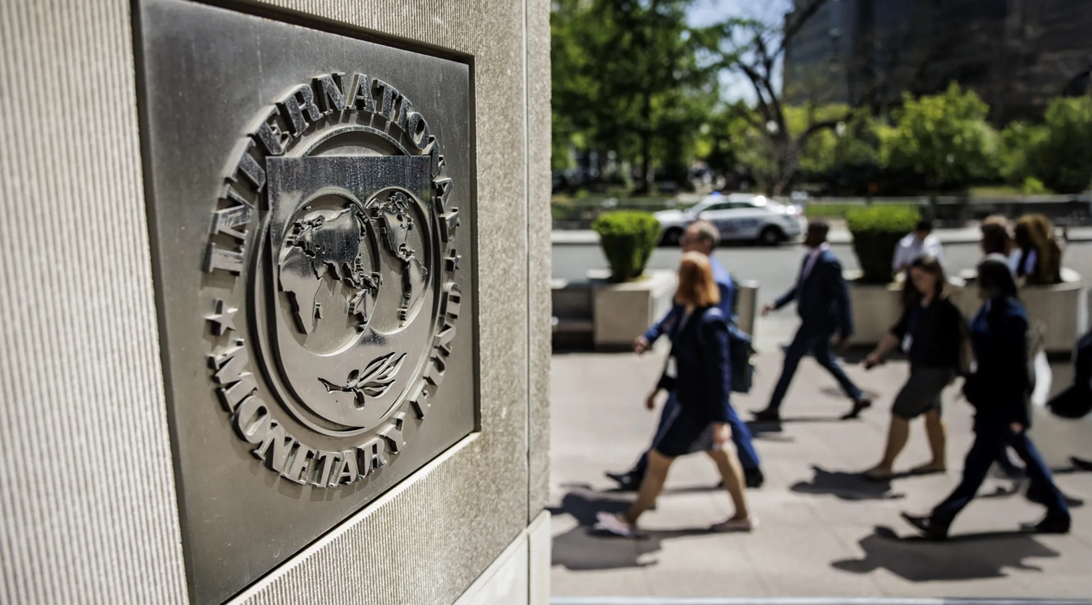 Placa metálica con el logotipo del Fondo Monetario Internacional en la entrada de un edificio, con personas caminando al fondo.