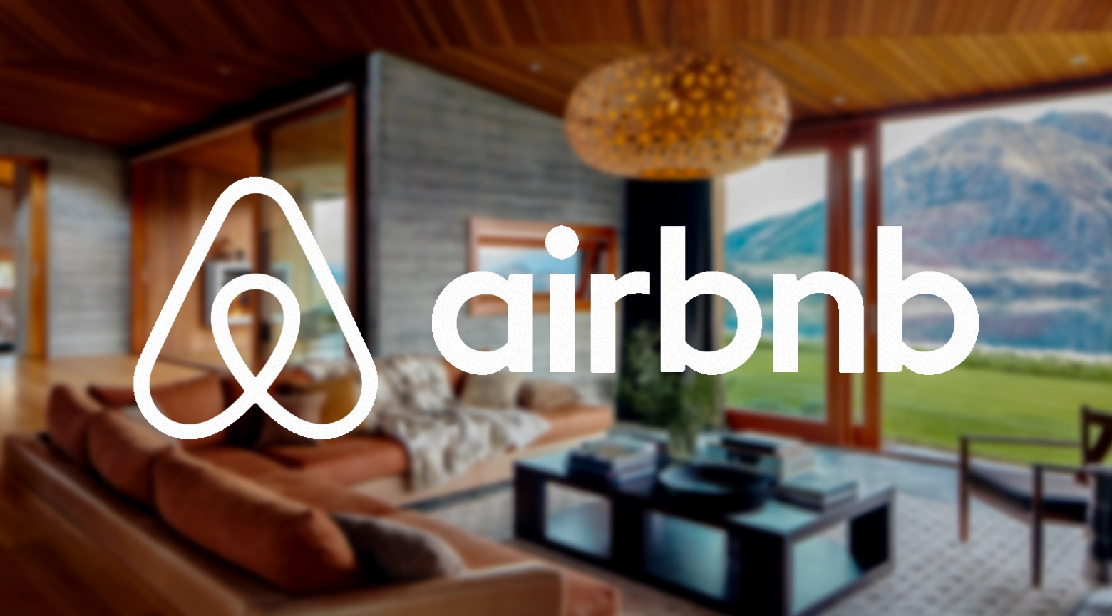 Logotipo de Airbnb en blanco sobre un fondo borroso de una sala moderna con grandes ventanales y vista a montañas y áreas verdes.