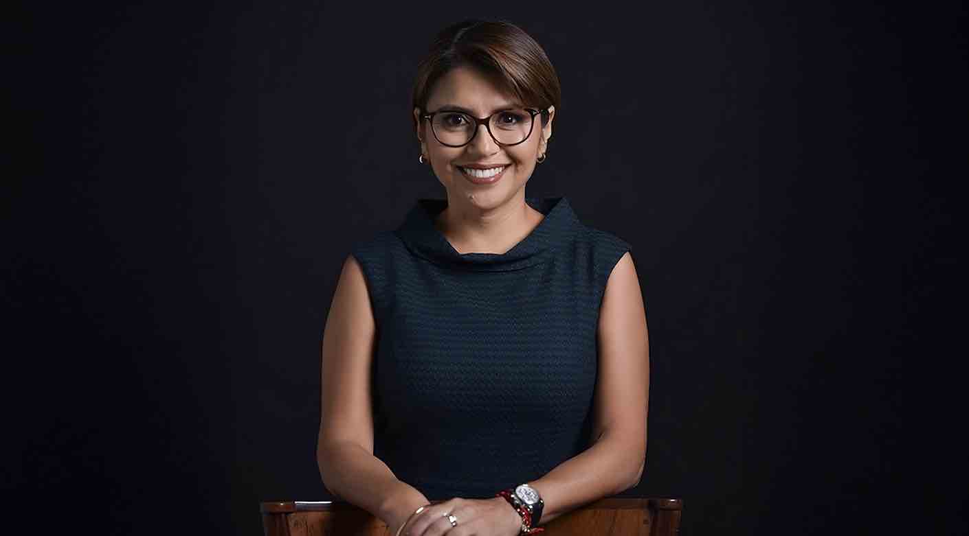 Retrato de una mujer sonriente con gafas, sentada frente a un fondo oscuro, en pose formal.