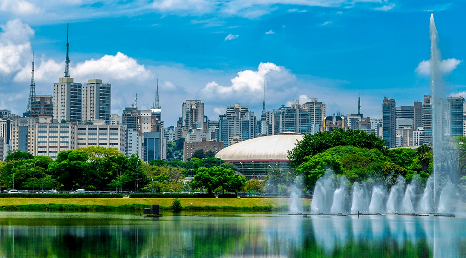 Vista panorámica de una gran ciudad moderna con edificios altos, vegetación abundante y fuentes sobre un lago en primer plano, bajo un cielo azul con nubes.