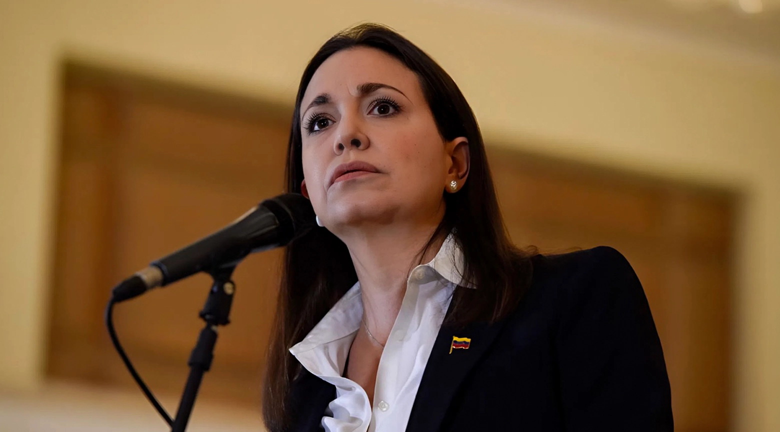 Mujer con chaqueta oscura y camisa blanca habla frente a un micrófono, con una pequeña bandera de Venezuela en la solapa.