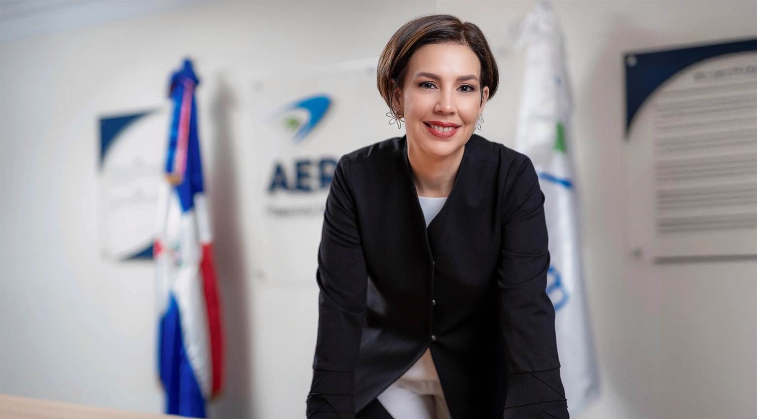 Mujer ejecutiva posando en una oficina institucional con banderas y logos en el fondo.