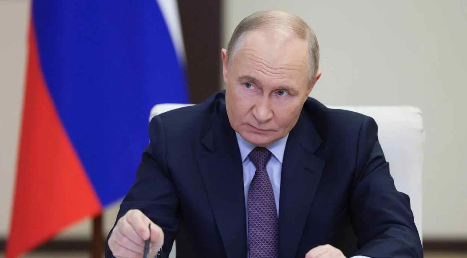 Vladimir Putin durante una reunión formal, con una expresión seria mientras firma un documento, con la bandera de Rusia al fondo.