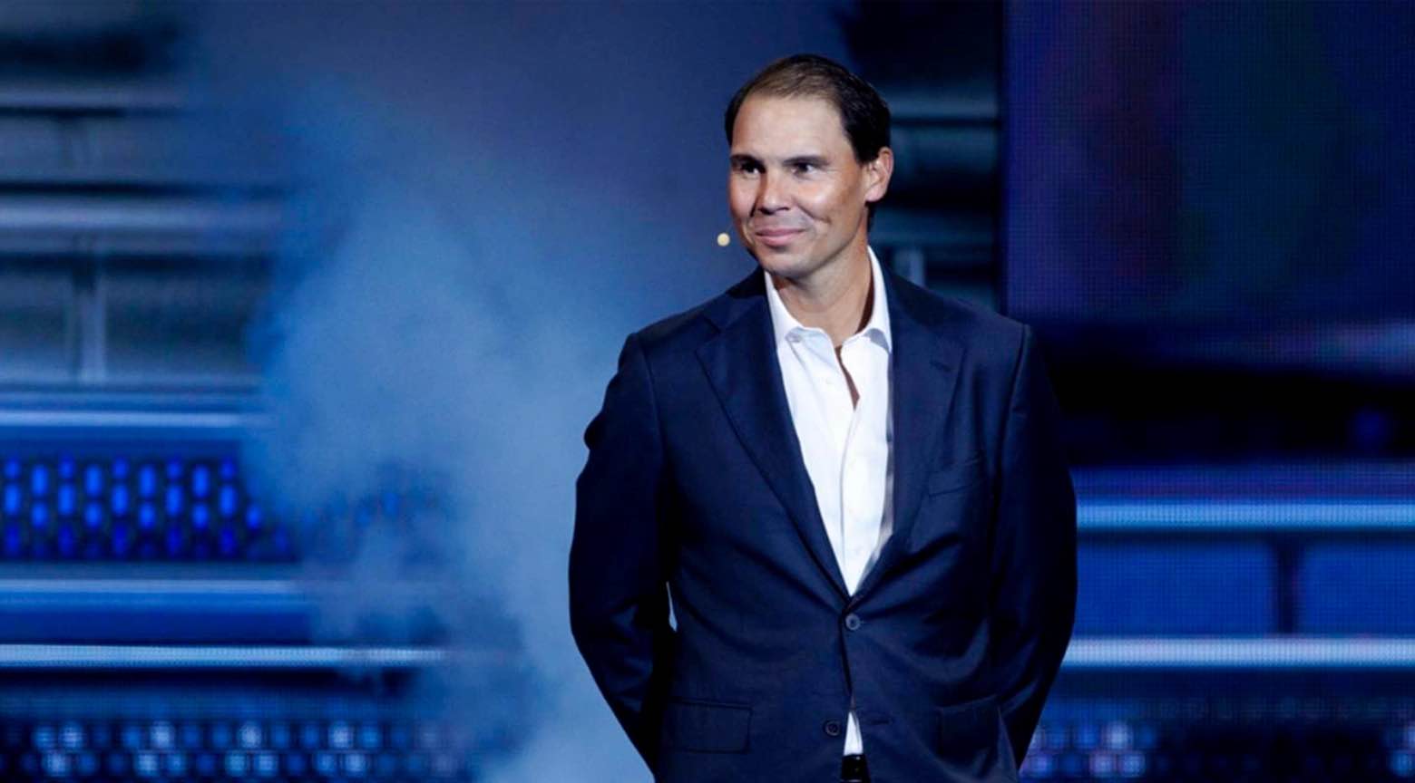 Rafael Nadal, vestido con traje oscuro y camisa blanca, aparece de pie sobre un escenario iluminado en tonos azules, con humo difuso a su alrededor.