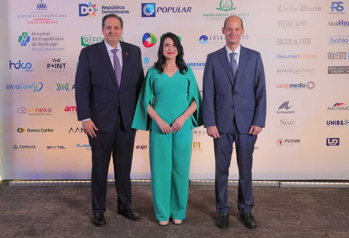 Víctor Atallah, Amelia Reyes Mora y Alejandro Cambiaso posan frente a un panel con los logos de las entidades patrocinadoras durante un evento sobre turismo de salud en República Dominicana.