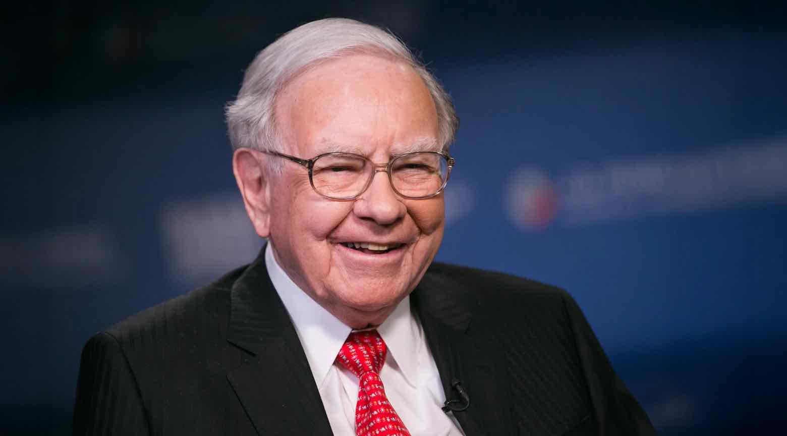 Warren Buffett sonríe mientras lleva puesto un traje negro y corbata roja con patrones, durante una entrevista.