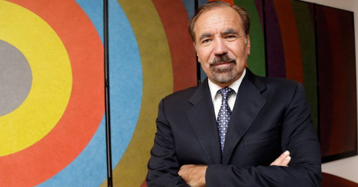 Jorge M. Pérez, empresario y filántropo, posa frente a una obra de arte colorida que destaca círculos de varios colores. Lleva un traje oscuro con una camisa y corbata de tonos claros.