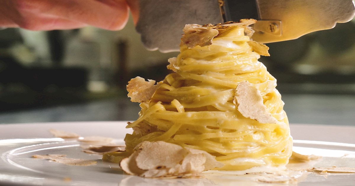 Nido de pasta fresca amarilla servido en forma vertical, coronado con láminas finas de trufa blanca sobre un plato blanco.