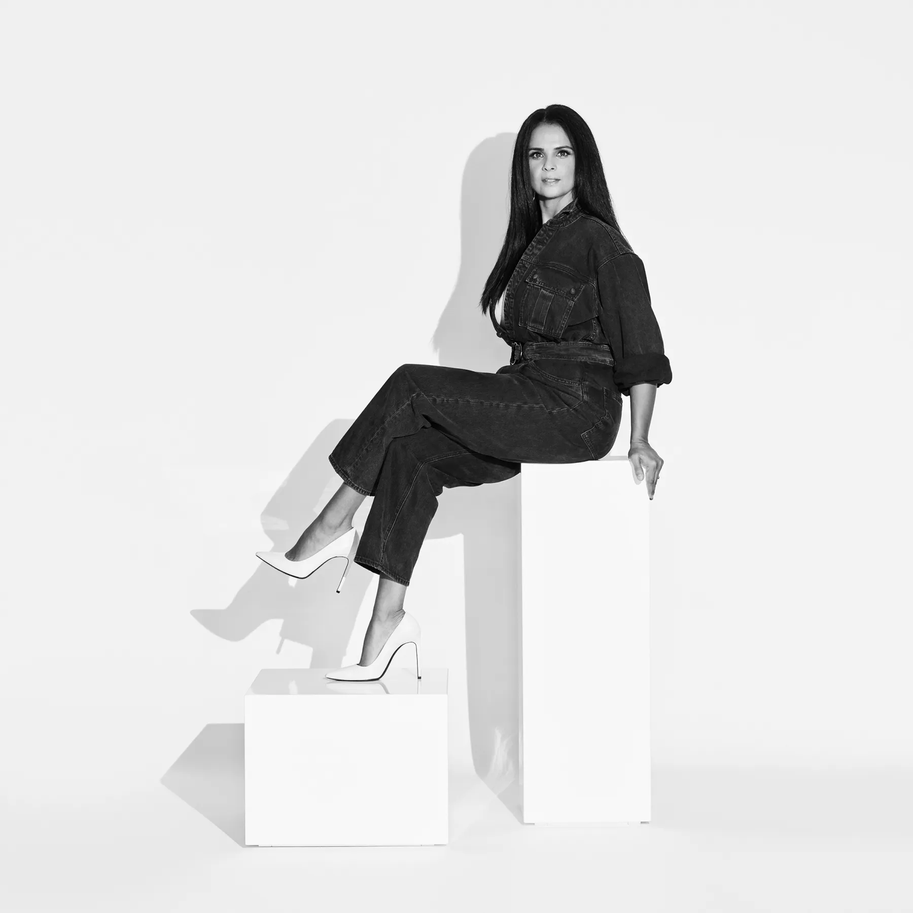 Retrato en blanco y negro de una mujer sentada sobre pedestales geométricos blancos, vestida con un enterizo oscuro y tacones claros, en una composición minimalista de estudio.