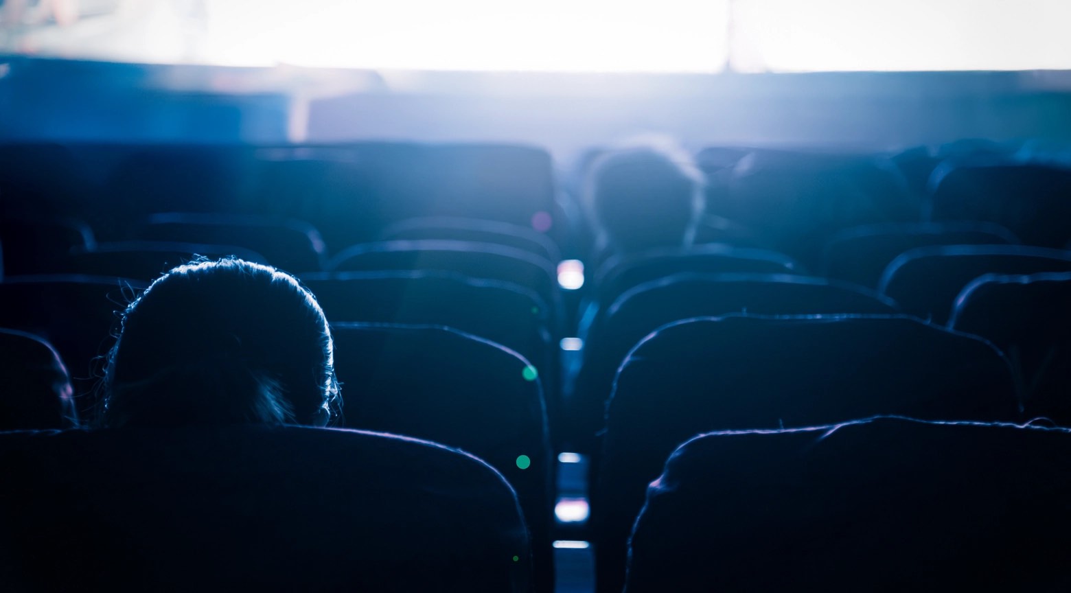 Sala de cine con butacas mayormente vacías y espectadores aislados, iluminada por la luz de la pantalla, reflejando la caída sostenida de la asistencia al cine.