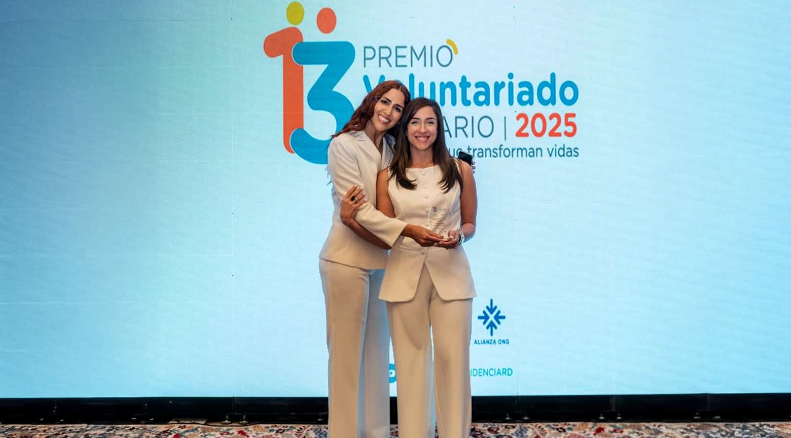 Dos mujeres vestidas de blanco posan sonrientes en el escenario durante la ceremonia del Premio Voluntariado Solidario 2025, sosteniendo un reconocimiento frente a una pantalla institucional.