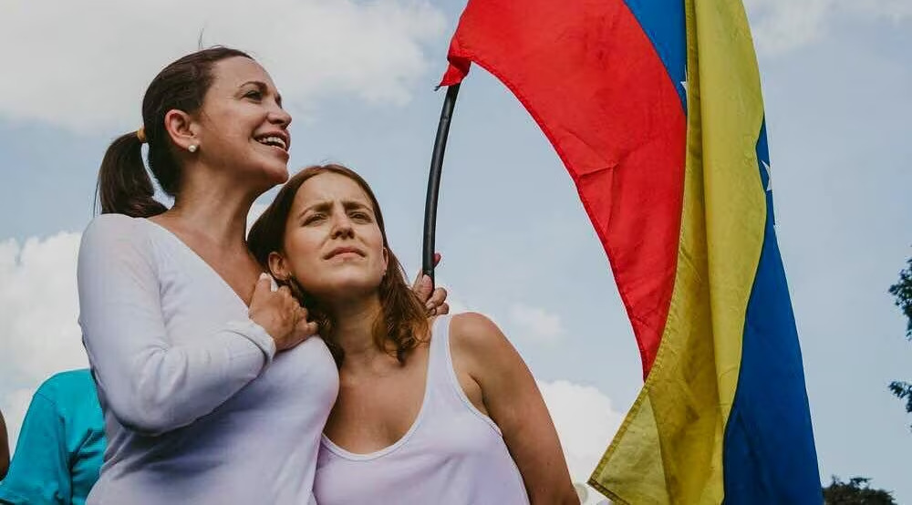 Dos mujeres de pie, abrazadas, sosteniendo una bandera de Venezuela. Ambas lucen sonrientes y se observan hacia el frente, mientras la bandera ondea en el viento.