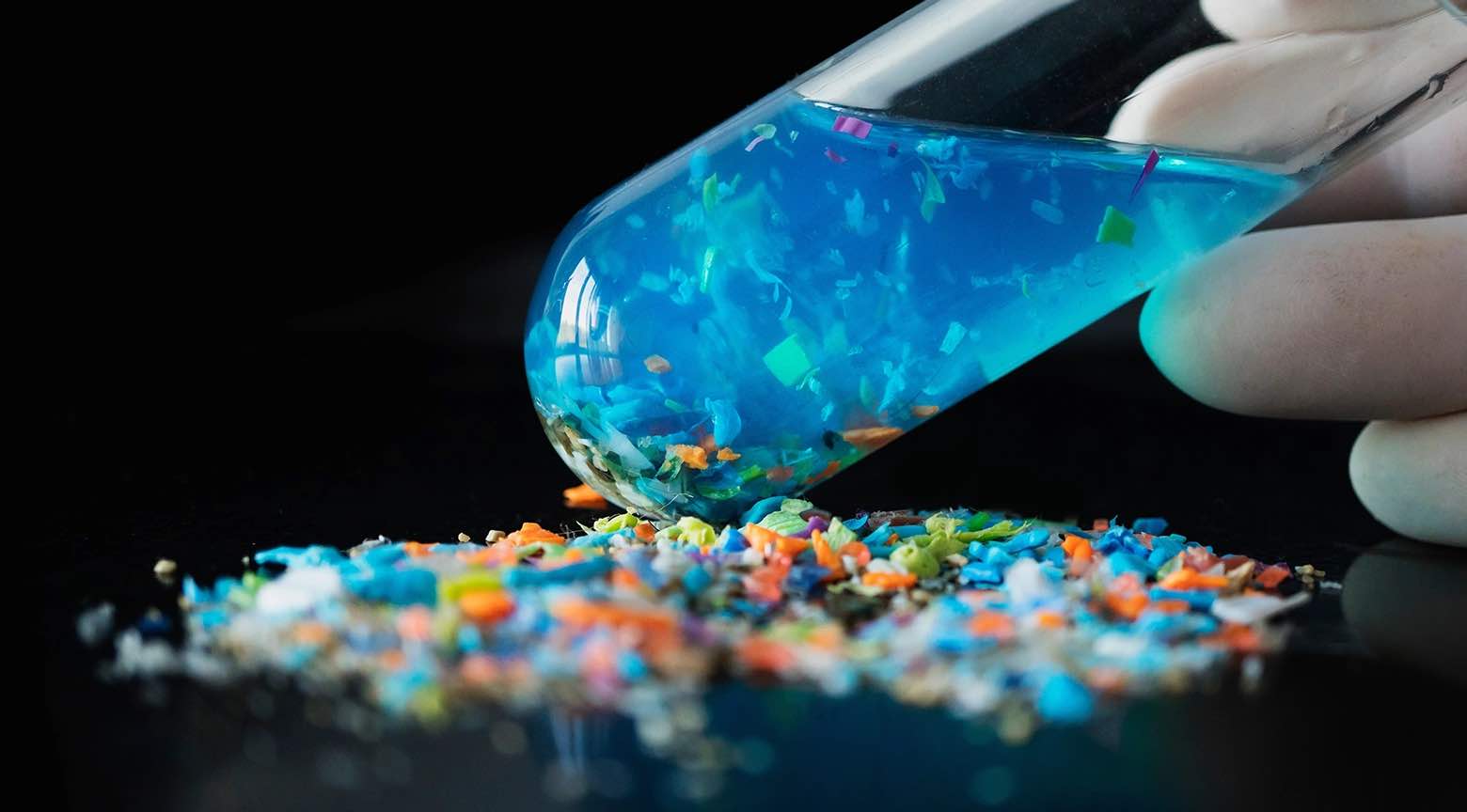 Tubo de ensayo con líquido azul y fragmentos de microplásticos dispersos sobre una superficie negra.