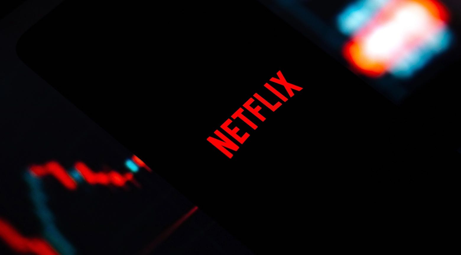 Logo de Netflix en rojo sobre una pantalla oscura, con gráficos bursátiles en tonos rojos y azules desenfocados en el fondo.
