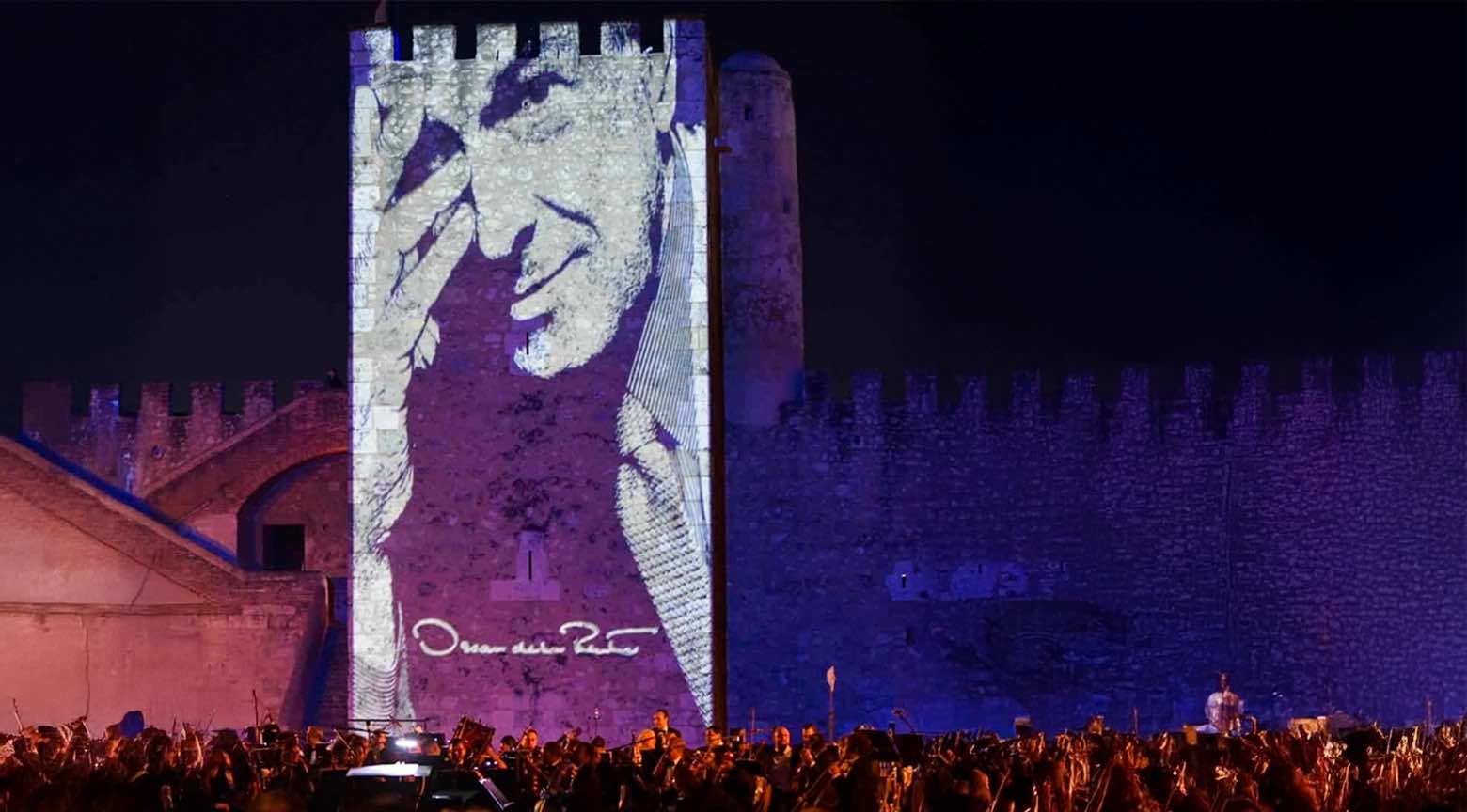 Proyección de una imagen en homenaje a Óscar de la Renta en una pared de un castillo, con una multitud observando.