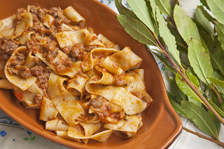 Plato de pappardelle con salsa de ragú de carne, servido en una fuente de barro, acompañado de hojas de laurel frescas.