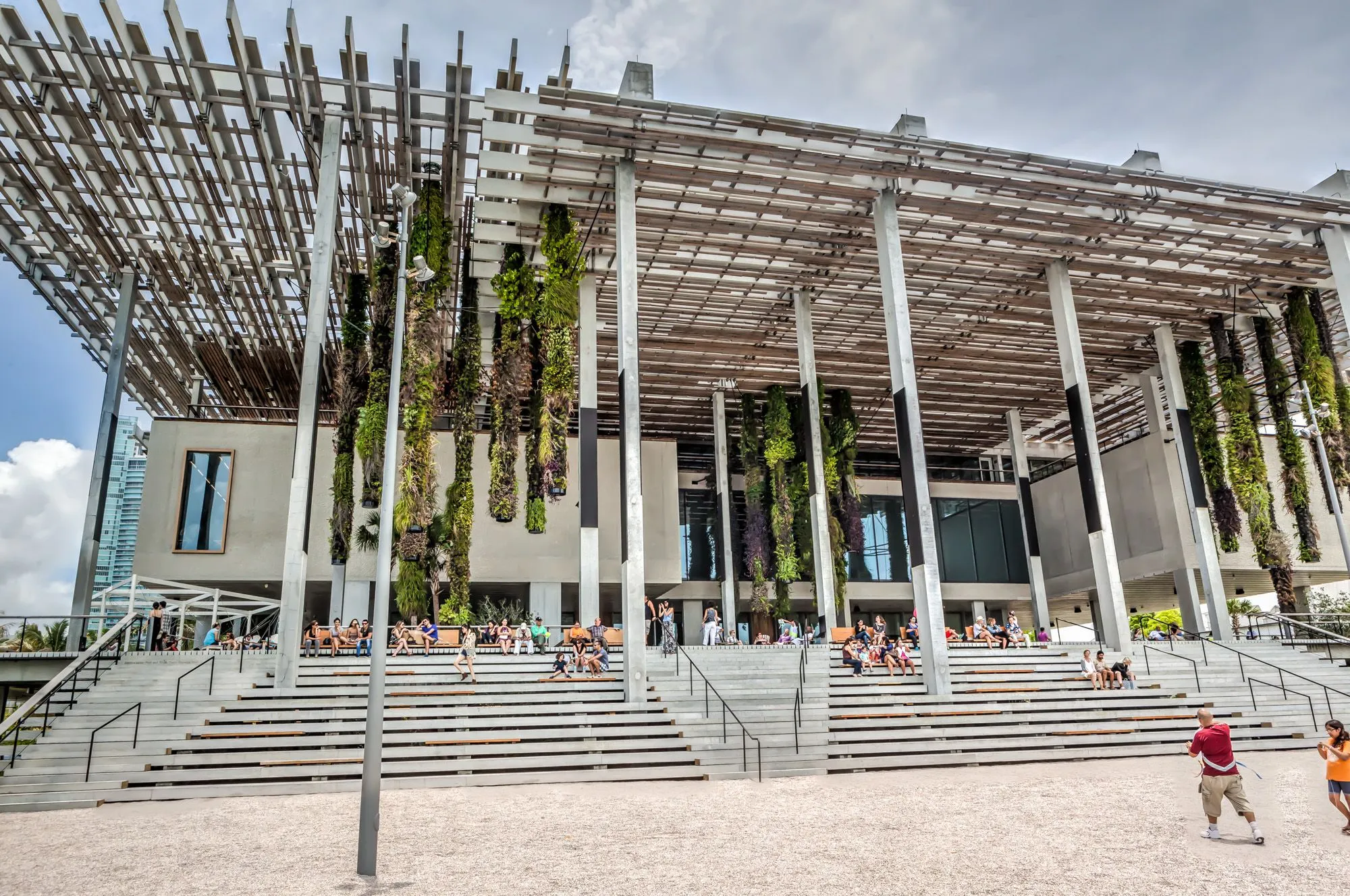 Vista exterior del Pérez Art Museum Miami (PAMM), con su arquitectura moderna y sostenible. Se observan las escaleras exteriores donde varias personas descansan, y la estructura del edificio se destaca por sus columnas y una pérgola con vegetación colgante.