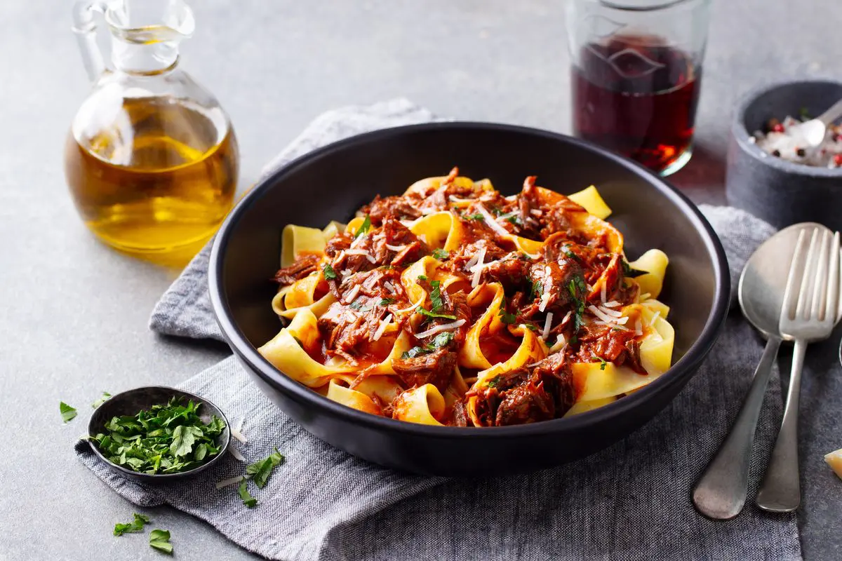 Plato de pappardelle con ragú de carne servido en un cuenco negro, cubierto con salsa espesa, hierbas frescas y queso rallado, acompañado de aceite de oliva y vino tinto.