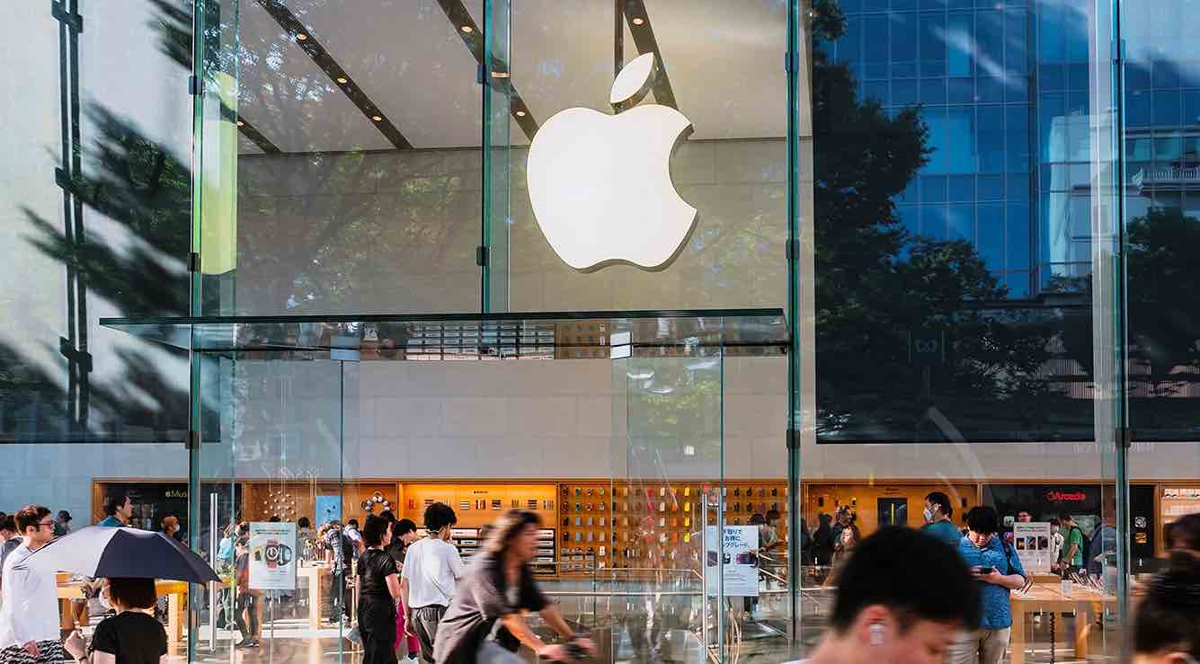Fachada de una Apple Store con logotipo blanco sobre una estructura de vidrio, mientras clientes y transeúntes se mueven frente al edificio.