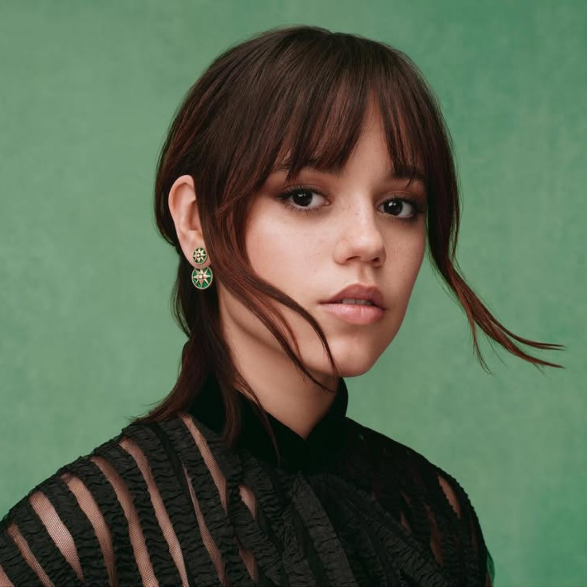 Retrato de una mujer joven con flequillo y cabello castaño recogido, vestida con un top negro de transparencias y textura, posando frente a un fondo verde mate con una expresión serena.