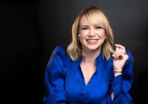 Mujer sonriente con blusa azul posa frente a un fondo oscuro, en un retrato de estudio de estilo profesional.