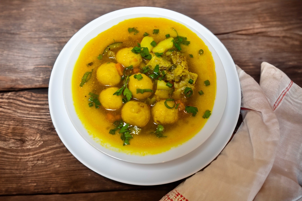 Sopa amarilla tradicional con bolitas de masa, trozos de carne y papas, servida caliente y decorada con hierbas frescas picadas.