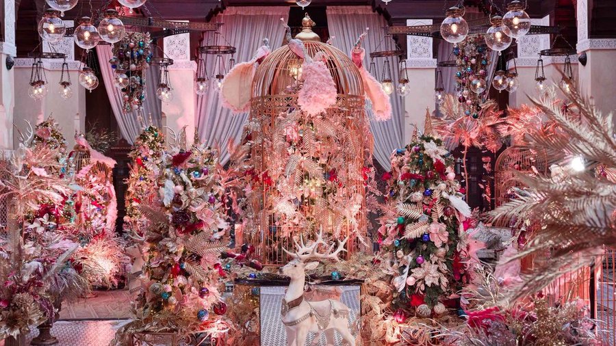 Escenografía navideña de lujo con árboles decorados en tonos rosados, dorados y metálicos, jaulas ornamentales, luces colgantes y detalles florales en un interior elegante y teatral.