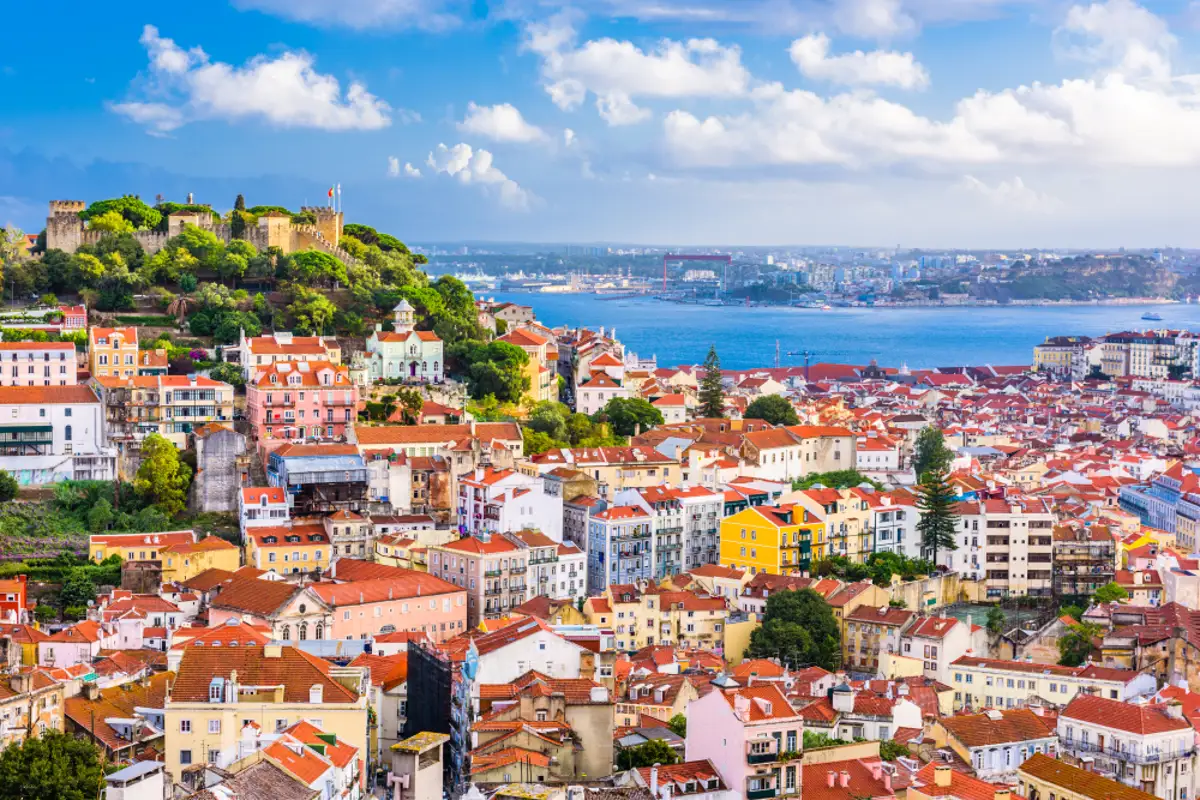 Vista panorámica del barrio de Alfama, en Lisboa, con tejados rojos, casas coloridas y el río Tajo al fondo bajo un cielo despejado.