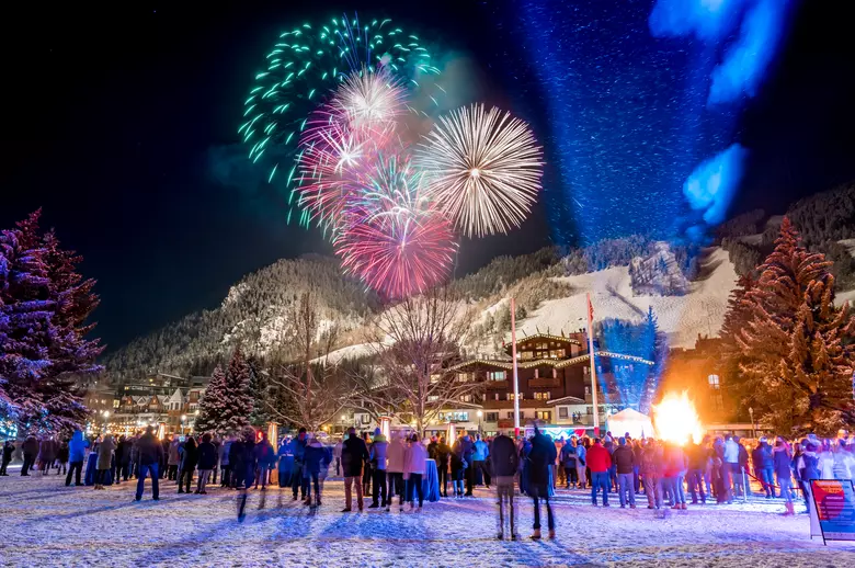 Espectáculo de fuegos artificiales sobre un pueblo alpino cubierto de nieve durante la celebración de Año Nuevo, con personas reunidas en una plaza invernal.