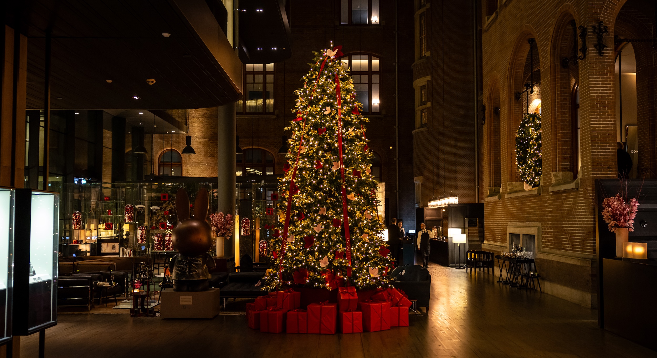 Árbol de Navidad de gran tamaño decorado con luces cálidas, lazos rojos y regalos en su base, ubicado en el elegante lobby de un hotel histórico con arquitectura de ladrillo y ambiente nocturno.