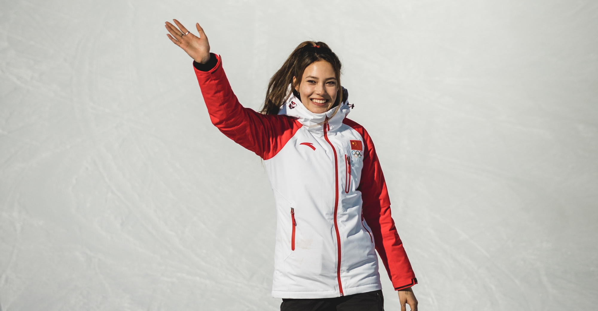 Atleta olímpica sonríe y saluda mientras viste chaqueta deportiva blanca y roja con emblema del equipo chino, sobre un fondo claro.