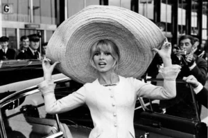 Mujer con vestido claro y un gran sombrero de ala ancha posa frente a fotógrafos en una escena urbana de los años sesenta.