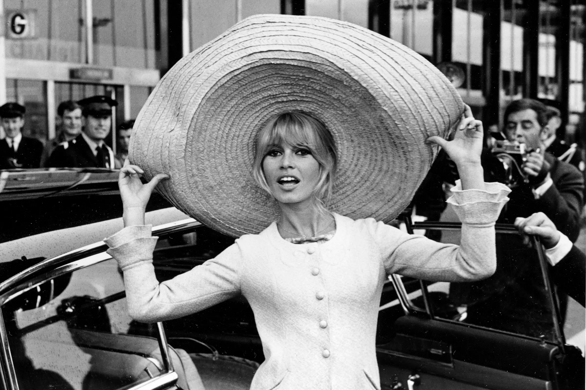 Mujer con vestido claro y un gran sombrero de ala ancha posa frente a fotógrafos en una escena urbana de los años sesenta.