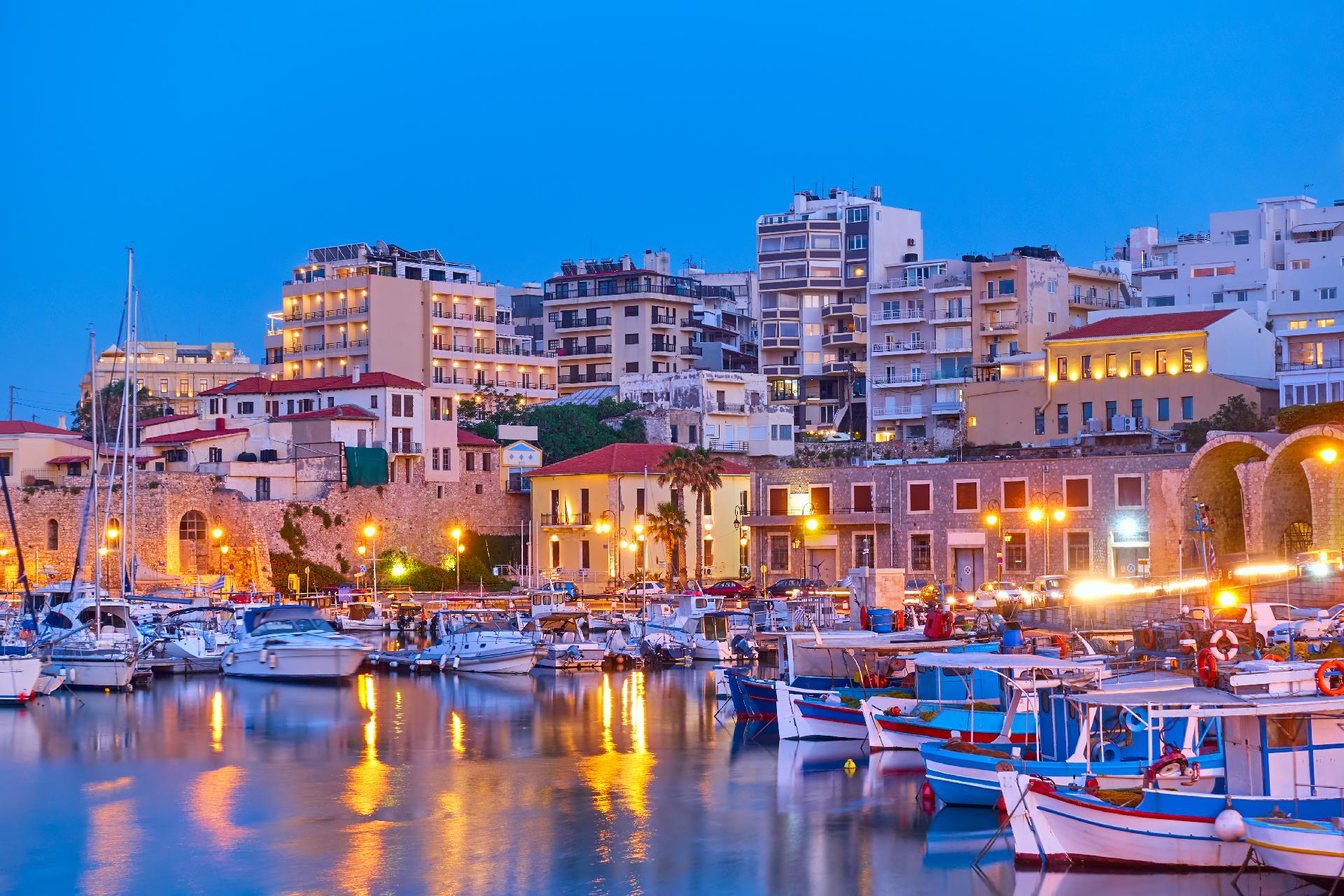 Puerto de Heraclión, en Creta, al atardecer, con barcos pesqueros amarrados y edificios iluminados reflejándose en el agua.