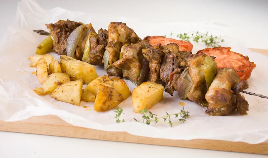 Kontosouvli servido en una brocheta, con trozos de carne asada intercalados con cebolla y pimiento, acompañado de papas asadas y tomate a la parrilla.