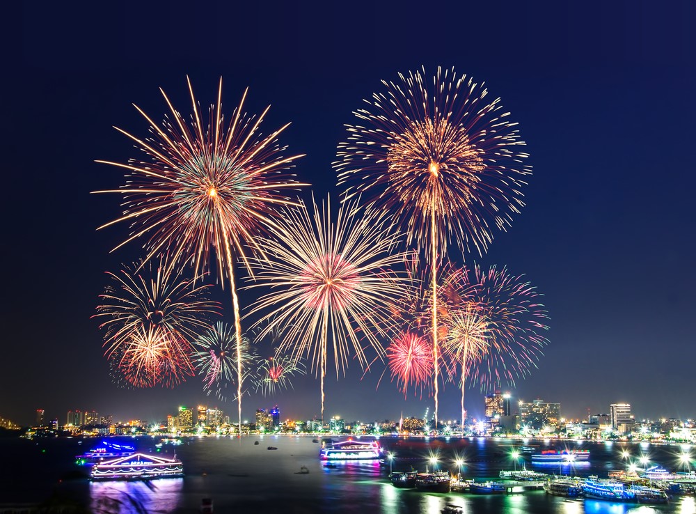 Fuegos artificiales iluminan el cielo nocturno sobre una ciudad costera durante la celebración de Año Nuevo, con embarcaciones navegando en primer plano.