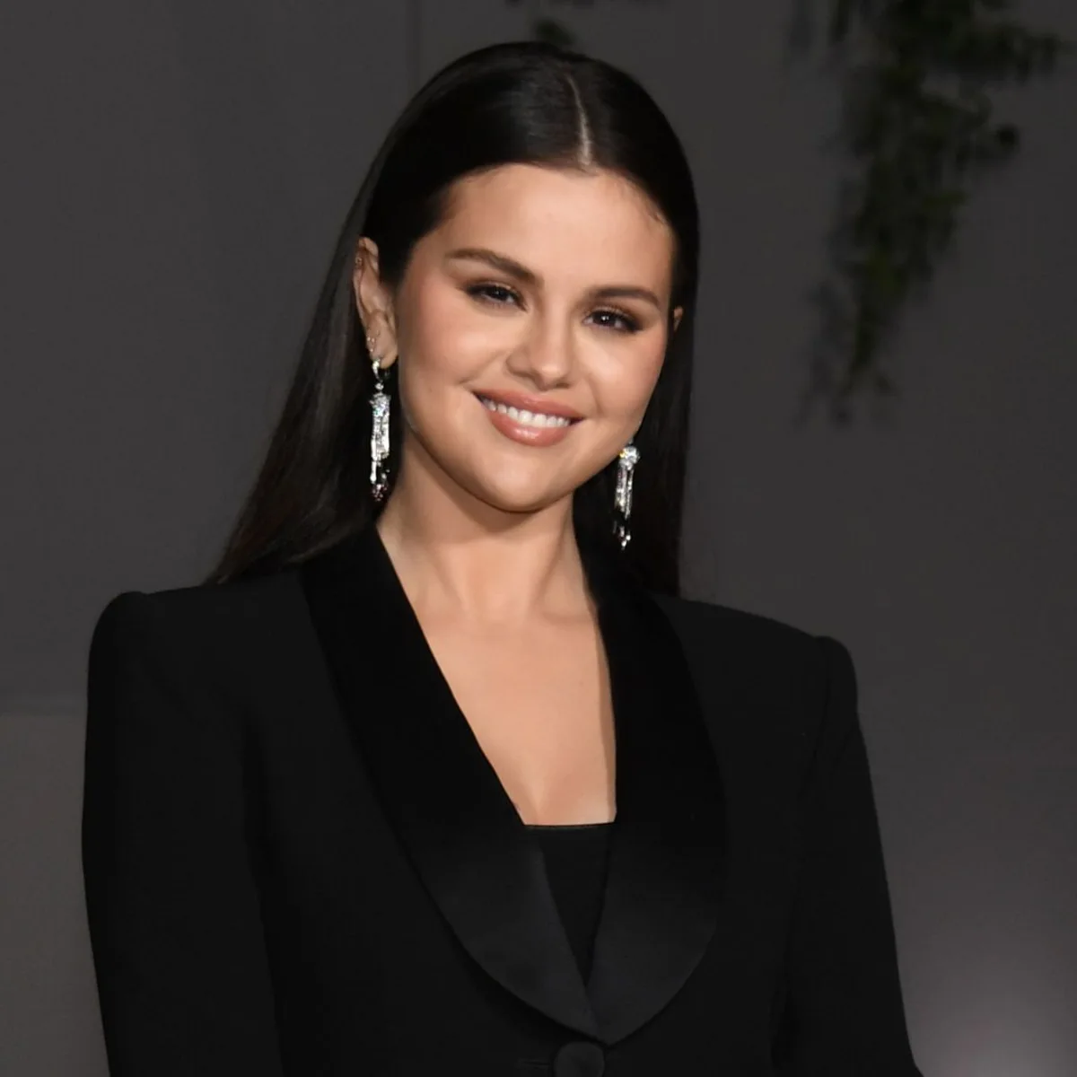 Mujer con cabello lacio oscuro y maquillaje natural posa sonriente frente a cámara, vestida con un elegante traje negro de corte clásico y pendientes largos brillantes, en un entorno de alfombra roja.