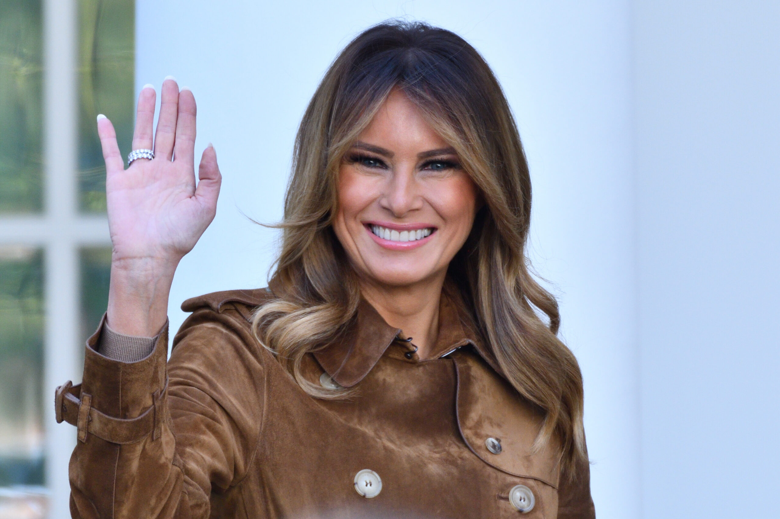 Mujer sonriente saluda levantando la mano, vestida con una chaqueta marrón, en un entorno exterior de estilo institucional.