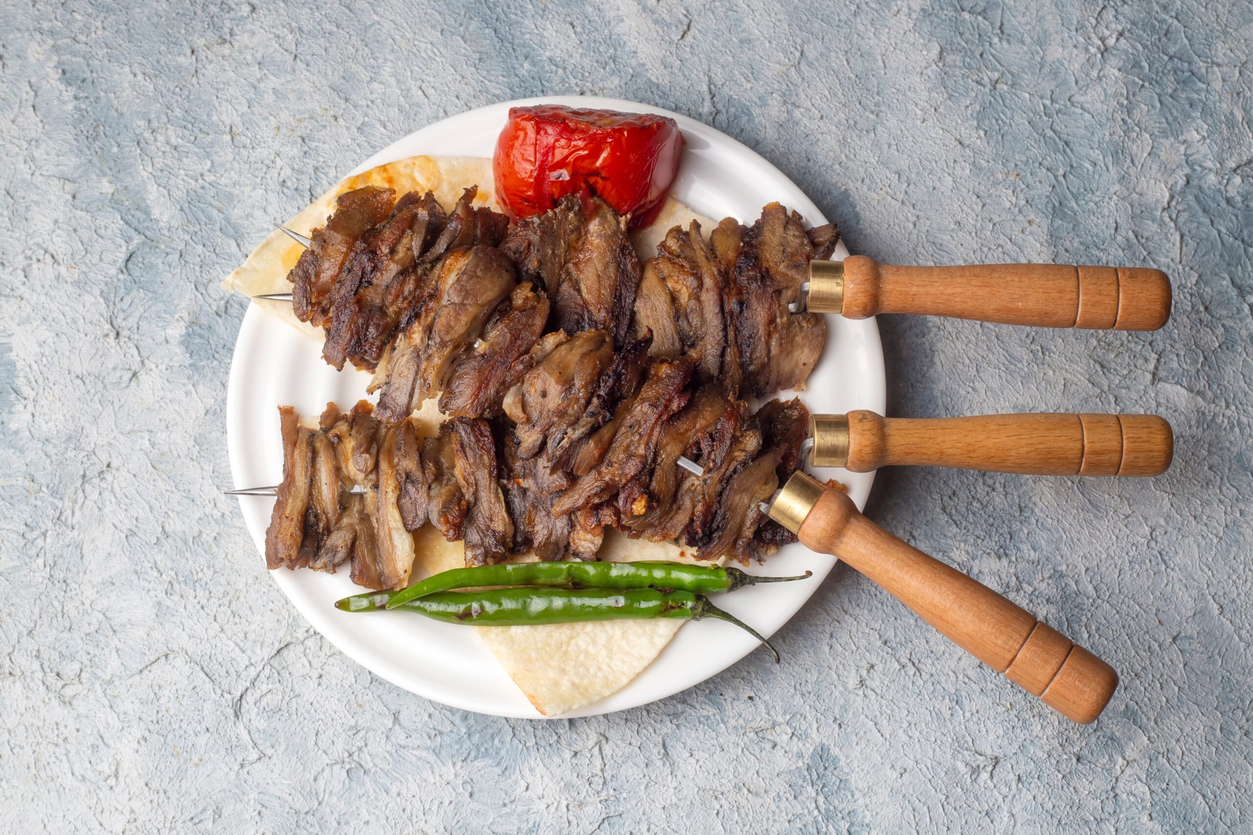 Brochetas de carne asada servidas sobre pan plano, acompañadas de tomate asado y pimientos verdes, presentadas en un plato blanco.