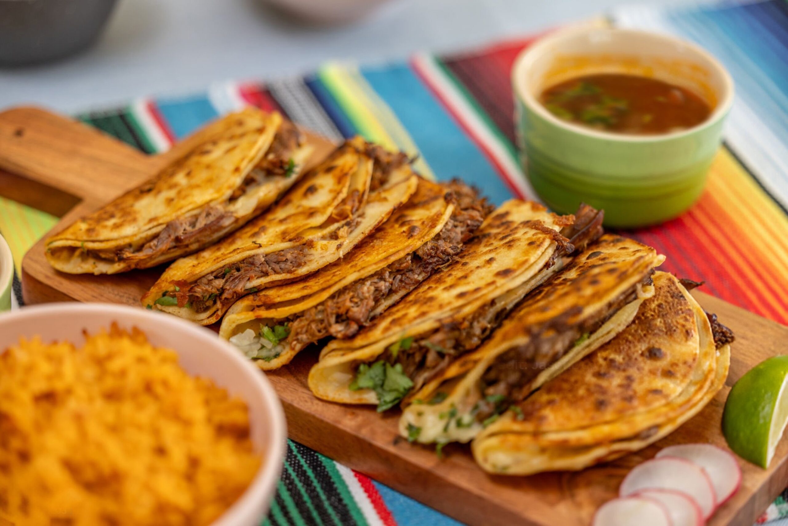 Quesabirrias doradas rellenas de carne, servidas en una tabla de madera, acompañadas de consomé, arroz, limón y rábanos.