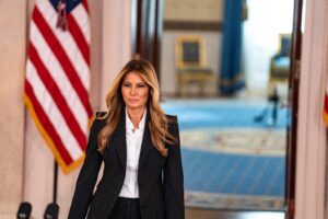 Mujer vestida con traje negro y camisa blanca camina por un pasillo institucional, con banderas de Estados Unidos al fondo, en un entorno oficial.